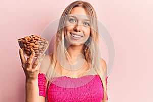 Young beautiful blonde woman holding bowl with almonds looking positive and happy standing and smiling with a confident smile