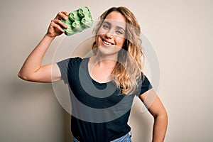 Young beautiful blonde woman holding box with fresh eggs over isolated white background with a happy face standing and smiling