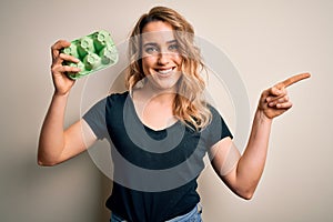 Young beautiful blonde woman holding box with fresh eggs over  white background very happy pointing with hand and finger