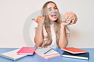 Young beautiful blonde woman holding brain while studying for school pointing thumb up to the side smiling happy with open mouth