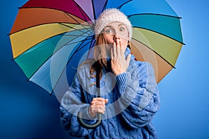 Young beautiful blonde woman holding colorful umbrella over isolated blue background cover mouth with hand shocked with shame for