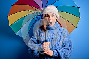 Young beautiful blonde woman holding colorful umbrella over isolated blue background scared in shock with a surprise face, afraid