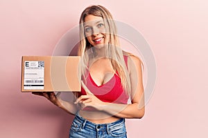 Young beautiful blonde woman holding delivery package looking positive and happy standing and smiling with a confident smile