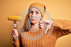 Young beautiful blonde woman holding fork with cobcorn over isolated yellow background with angry face, negative sign showing