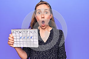 Young beautiful blonde woman holding holidays trip calendar scared and amazed with open mouth for surprise, disbelief face