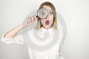 Young beautiful blonde woman holding magnifying glass near her eye looks surprised isolated white background