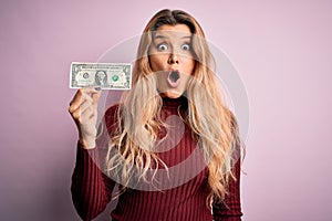 Young beautiful blonde woman holding one dollar banknote over isolated pink background scared in shock with a surprise face,