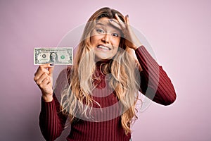 Young beautiful blonde woman holding one dollar banknote over isolated pink background stressed with hand on head, shocked with