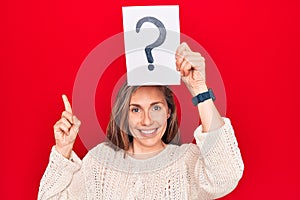 Young beautiful blonde woman holding question mark over head smiling happy pointing with hand and finger to the side