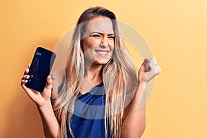 Young beautiful blonde woman holding smartphone showing screen over yellow background screaming proud, celebrating victory and