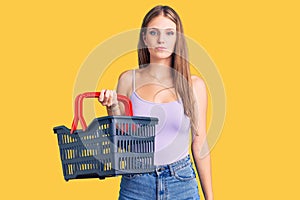 Young beautiful blonde woman holding supermarket shopping basket thinking attitude and sober expression looking self confident