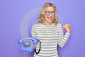 Young beautiful blonde woman holding vintage telephone over isolated purple background pointing thumb up to the side smiling happy