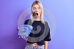 Young beautiful blonde woman holding vintage telephone over isolated purple background scared and amazed with open mouth for