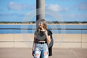 Young, beautiful blonde woman leaning against a lamppost. The woman is dressed in casual clothes and wears a black leather jacket