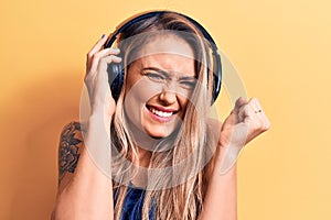 Young beautiful blonde woman listening to music using headphones over yellow background screaming proud, celebrating victory and