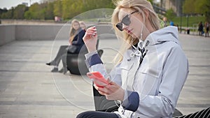 A young beautiful blonde woman listens to music on her smartphone using headphones. A young woman enjoys a walk along