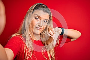 Young beautiful blonde woman making selfie by the camer over isolated red background with angry face, negative sign showing