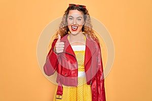 Young beautiful blonde woman pin-up with blue eyes wearing red sunglasses and jacket doing happy thumbs up gesture with hand