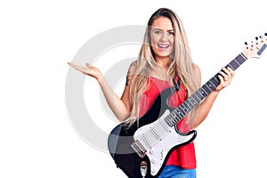 Young beautiful blonde woman playing electric guitar celebrating victory with happy smile and winner expression with raised hands