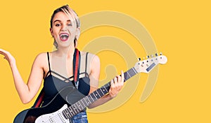 Young beautiful blonde woman playing electric guitar celebrating victory with happy smile and winner expression with raised hands