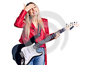 Young beautiful blonde woman playing electric guitar stressed and frustrated with hand on head, surprised and angry face