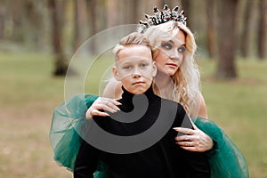 Young beautiful blonde woman queen with young boy in black outfit. Princess mother walks with son. autumn green forest mystic.