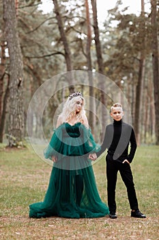 Young beautiful blonde woman queen with young boy in black outfit. Princess mother walks with son. autumn green forest mystic.