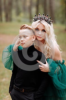 Young beautiful blonde woman queen with young boy in black outfit. Princess mother walks with son. autumn green forest mystic.