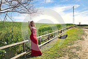 Young, beautiful, blonde woman with a red dress and flower tiara, eyes closed, leaning on a wooden railing with a meadow in the