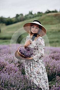 Young beautiful blonde woman in romantic dress, straw hat and basket of flowers posing in lavender field. Soft selective focus