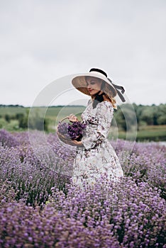 Young beautiful blonde woman in romantic dress, straw hat and basket of flowers posing in lavender field. Soft selective focus