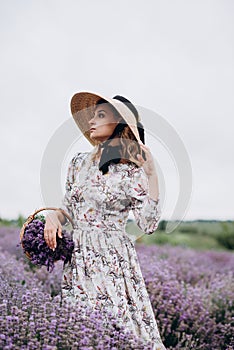 Young beautiful blonde woman in romantic dress, straw hat and basket of flowers posing in lavender field. Soft selective focus