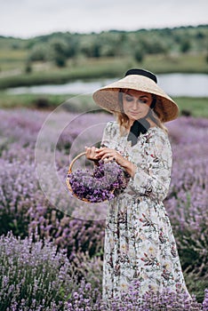 Young beautiful blonde woman in romantic dress, straw hat and basket of flowers posing in lavender field. Soft selective focus