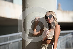 A young, beautiful blonde woman with sunglasses, wearing a crochet top and baggy pants, leaning between a column and a railing