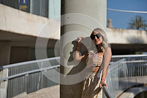 A young, beautiful blonde woman with sunglasses, wearing a crochet top and baggy pants, leaning between a column and a railing