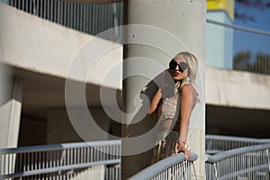 A young, beautiful blonde woman with sunglasses, wearing a crochet top and baggy pants, leaning between a column and a railing