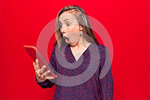 Young beautiful blonde woman using tablet over isolated red background scared and amazed with open mouth for surprise, disbelief
