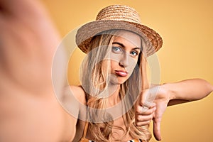 Young beautiful blonde woman on vacation wearing bikini and hat making selfie by camera with angry face, negative sign showing