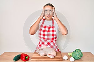 Young beautiful blonde woman wearing apron cooking chicken with hand on head, headache because stress