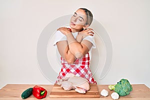 Young beautiful blonde woman wearing apron cooking chicken hugging oneself happy and positive, smiling confident