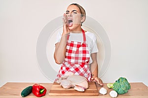 Young beautiful blonde woman wearing apron cooking chicken shouting and screaming loud to side with hand on mouth