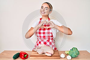 Young beautiful blonde woman wearing apron cooking chicken smiling in love doing heart symbol shape with hands