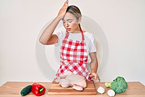 Young beautiful blonde woman wearing apron cooking chicken surprised with hand on head for mistake, remember error