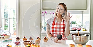 Young beautiful blonde woman wearing apron cooking pastries looking for recipe on smartphone doing ok sign with fingers, smiling