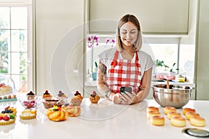 Young beautiful blonde woman wearing apron cooking pastries looking for recipe on smartphone looking positive and happy standing