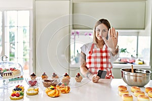 Young beautiful blonde woman wearing apron cooking pastries looking for recipe on smartphone with open hand doing stop sign with