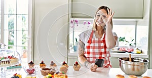 Young beautiful blonde woman wearing apron cooking pastries looking for recipe on smartphone smiling happy doing ok sign with hand
