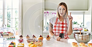Young beautiful blonde woman wearing apron cooking pastries looking for recipe on smartphone surprised with an idea or question