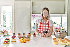 Young beautiful blonde woman wearing apron cooking pastries looking for recipe on smartphone thinking attitude and sober