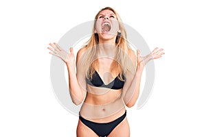 Young beautiful blonde woman wearing bikini crazy and mad shouting and yelling with aggressive expression and arms raised